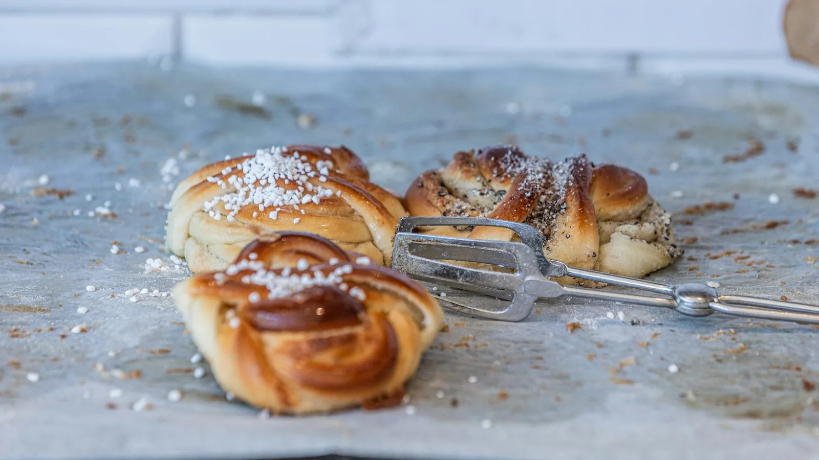 Tre kanelbullar med pärlsocker ligger på bakplåtspapper bredvid en elvisp